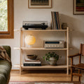 A light natural wood open shelving unit styled in a cozy living room setting, holding a turntable, records, a glowing globe lamp, books, and a small potted plant.