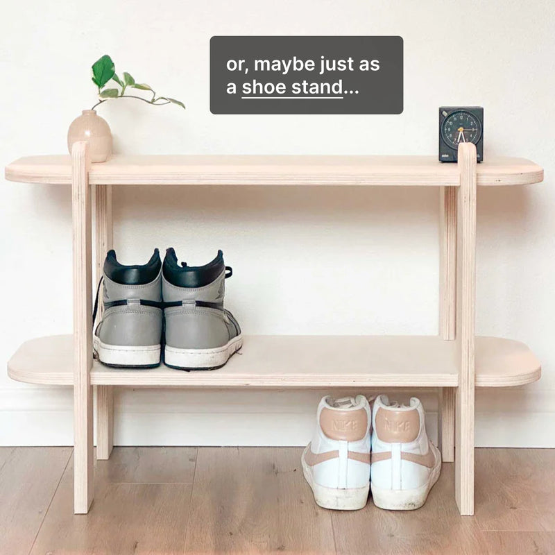 Minimalist shoe stand displaying two pairs of sneakers, with a potted plant and clock in the background.