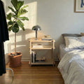 Modern Nightstand with books, lamp, and plant beside a cozy bed in a sunlit bedroom.